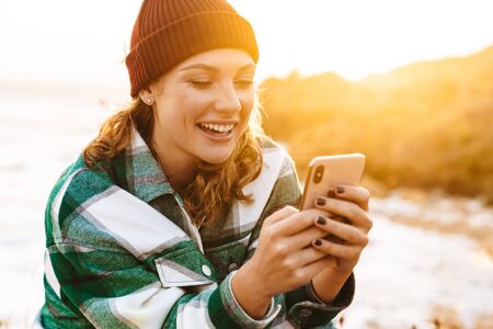 Image of caucasian joyful woman using smartphone and laughing while sitting at seaside