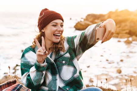 Image of joyful young caucasian woman wearing hat and plaid shirt taking selfie photo on cellphone while walking by seasideの写真素材