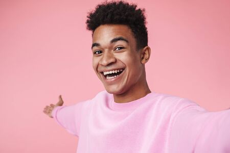 Portrait of a smiling african teenager boy wearing pullower standing isolated over pink background, taking a selfieの写真素材