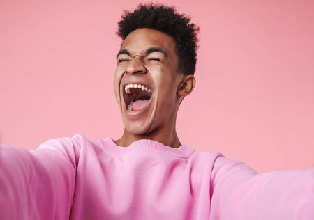 Portrait of a smiling african teenager boy wearing pullower standing isolated over pink background, taking a selfie, screamingの写真素材