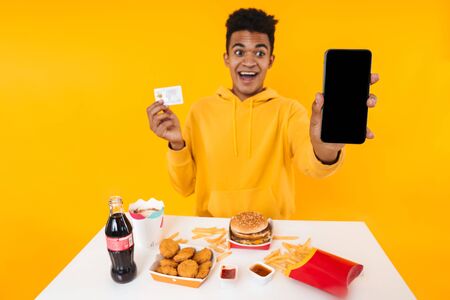Happy african teenager boy eating fast food meal while sitting at the table isolated over yellow background, showing blank screen mobile phone and credit cardの写真素材