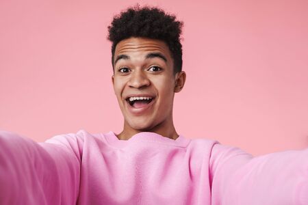 Portrait of a smiling african teenager boy wearing pullower standing isolated over pink background, taking a selfie, screamingの写真素材