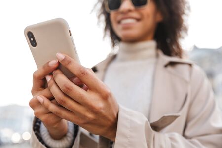 Smiling attractive young african woman wearing autumn coat walking outdoors, using mobile phoneの写真素材
