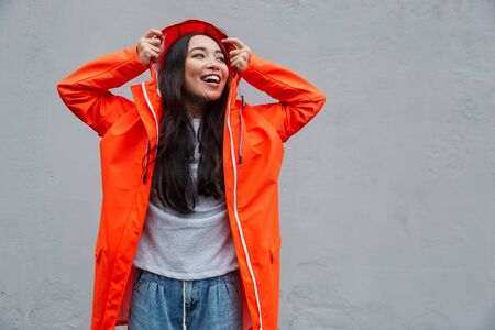 Smiling young asian woman wearing raincoat walking outdoors in the rain, posing in a hood