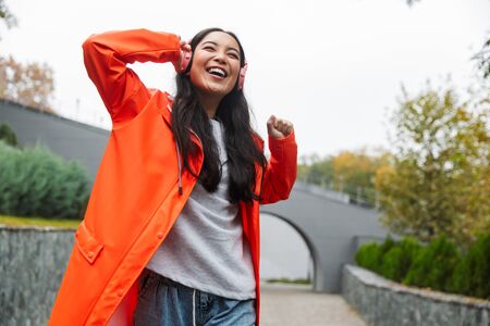 Smiling young asian woman wearing raincoat walking outdoors in the rain, listening to music with wireless headphonesの写真素材