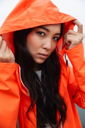 Attractive young asian woman wearing raincoat walking outdoors in the rain, posing in a hoodの写真素材