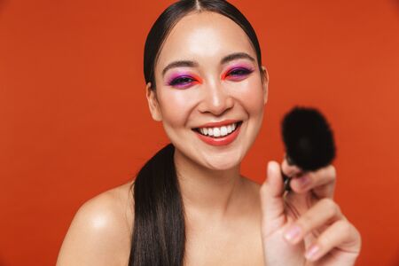 Beauty portrait of an attractive happy young topless asian woman with brunette hair wearing bright makeup, standing isolated over red background, holding makeup brushの写真素材