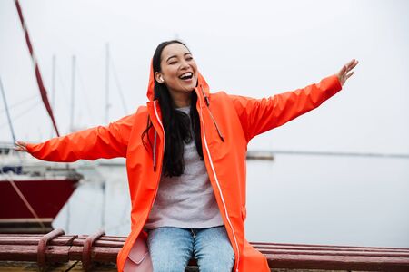 Smiling cheerful attractive young asian woman wearing raincoat spending time outdoors walking at the coastlandの写真素材
