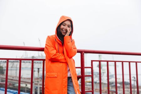 Smiling young asian woman wearing raincoat walking outdoors in the rain, posing in a hoodの写真素材