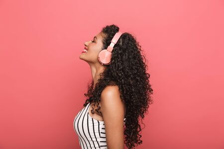 Image of happy african american woman with curly hair listening to music with headphones isolated over pink backgroundの写真素材