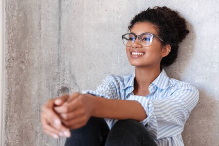 Beautiful smiling young african woman wearing casual clothes and glasses leaning on a wall indoors, posing while sittingの写真素材