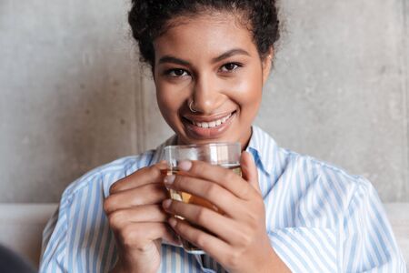 Smiling attractive young african woman wearing shirt relaxing on a couch at home, drinking cup of teaの写真素材