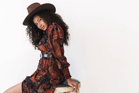 Beautiful cheerful young woman wearing cowboy hat standing while leaning on a chair isolated over...