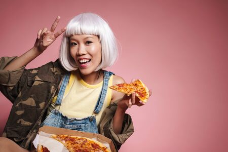 Image of young asian girl wearing white wig having fun and eating pizza isolated over pink backgroundの写真素材