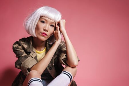 Image of young asian girl wearing white wig sitting and looking at camera isolated over pink backgroundの写真素材