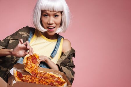 Image of young asian girl wearing white wig having fun and eating pizza isolated over pink backgroundの写真素材