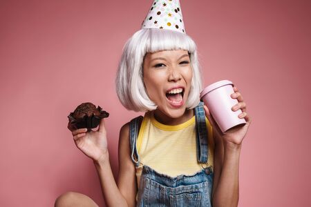 Image of young asian girl wearing party cone drinking coffee and eating cupcake isolated over pink backgroundの写真素材