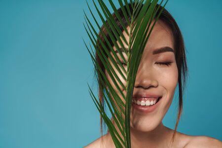 Photo of asian smiling woman posing  isolated over blue wall background holding plant leaf.の写真素材