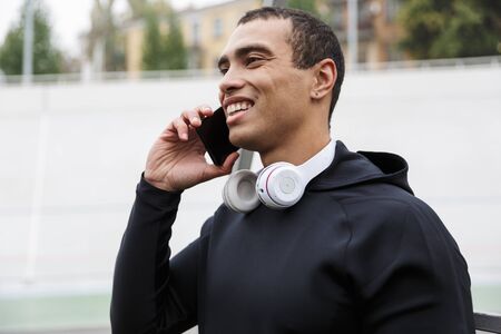 Photo of athletic smiling sportsman in tracksuit talking cellphone while working out on stadium...