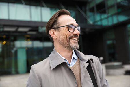 Image of handsome joyful businessman in eyeglasses posing and smiling near office building in urban...