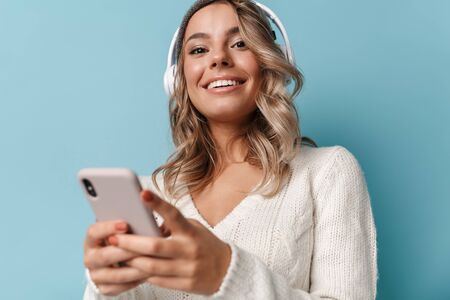 Portrait of cute smiling woman in knit hat using headphones and cellphone isolated over blue...