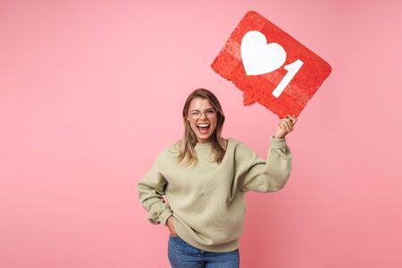 Portrait of laughing woman in eyeglasses holding placard and winking isolated over pink background