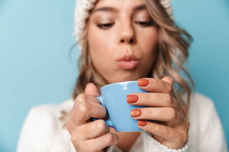 Portrait of blonde attractive woman in white hat blowing and holding cup isolated over blue backgroundの写真素材