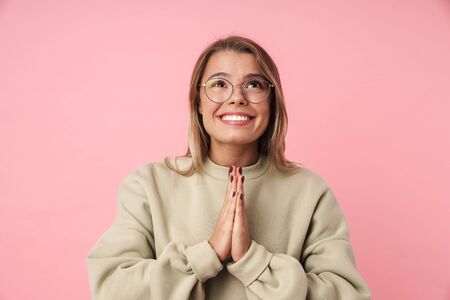 Portrait of young pleased woman in eyeglasses smiling and holding palms together isolated over pink...
