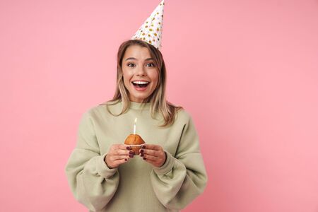 Portrait of excited happy woman in party cone holding cake and looking at camera isolated over pink...
