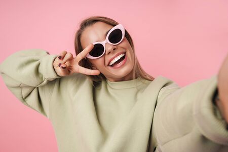Portrait of beautiful smiling woman in sunglasses gesturing peace sign and taking selfie isolated over pink backgroundの写真素材