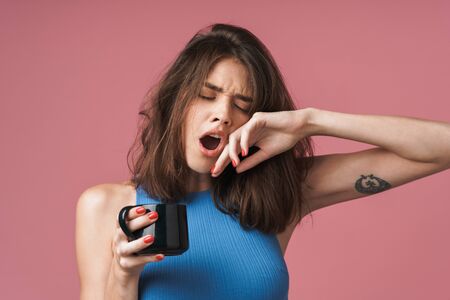 Portrait of a young tired attractive brunette woman standing isolated over pink background, holding...