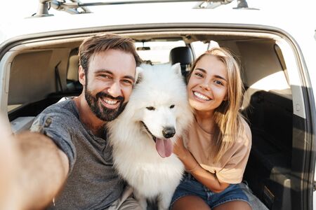 Cheerful young couple at the beach, playing with their dog, sitting inside a car, taking a selfieの写真素材