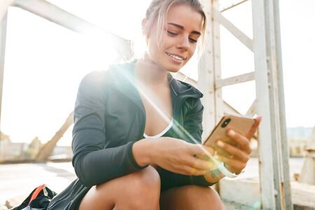 Beautiful smiling young sportsgirl sitting on a bridge, listening to music with earphones, holding mobile phoneの写真素材