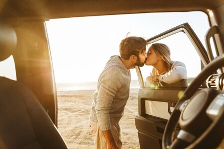 Photo of young happy cheerful loving couple outdoors at beach near car kissing.