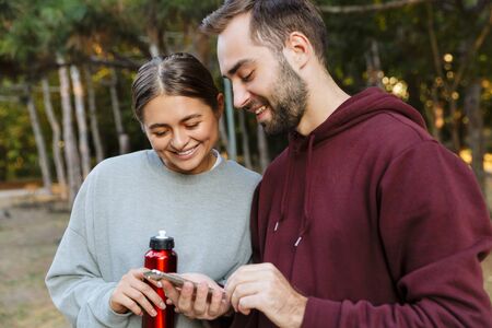 Photo of a young happy positive sports woman and man using mobile phone outdoors in nature green...