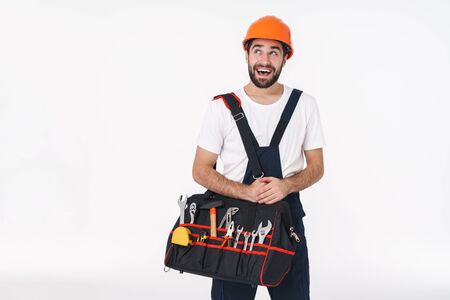 Image of excited smiling young man builder in helmet isolated over white wall background holding bag with equipment instruments looking aside.の写真素材