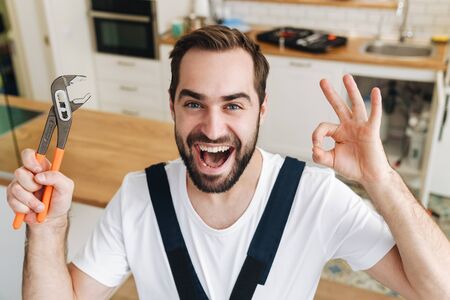 Image of young positive happy man plumber work in uniform indoors holding pipe wrench showing okay...