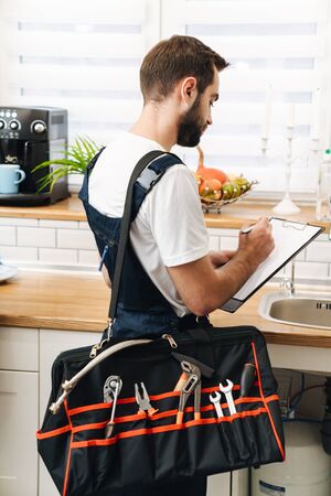 Image of young man plumber work in uniform indoors holding bag with equipment and clipboard.