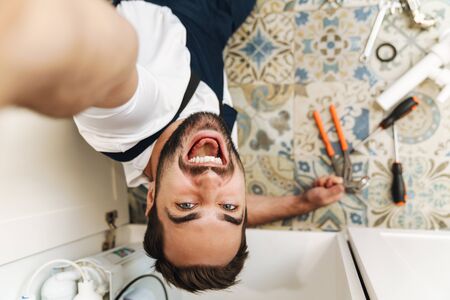 Image of a handsome cheery optimistic young man plumber work in uniform indoors take a selfie by camera.の写真素材