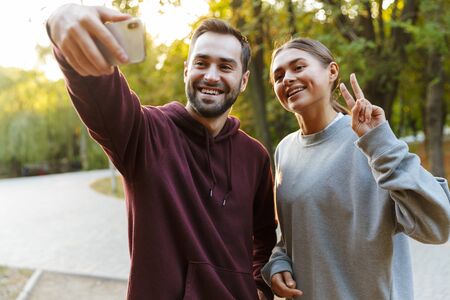 Photo of beautiful happy couple in sportswear taking selfie photo on cellphone and gesturing peace...
