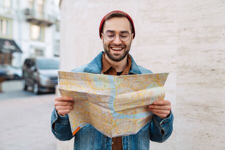 Close up of a handsome happy young stylish bearded man walking outdoors in the street, holding city guide mapの写真素材