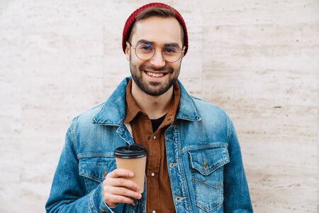 Close up of a handsome happy young stylish bearded man walking outdoors in the street, holding takeaway coffee cupの写真素材