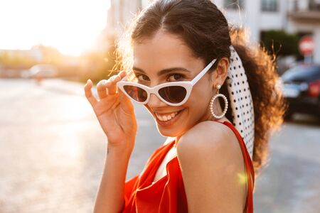 Image closeup of beautiful joyful woman in sunglasses looking at camera and smiling while walking on city streetの写真素材