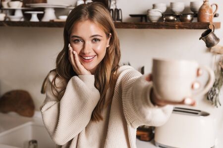 Image of beautiful funny woman wearing casual clothes drinking coffee and looking at camera in cozy kitchenの写真素材