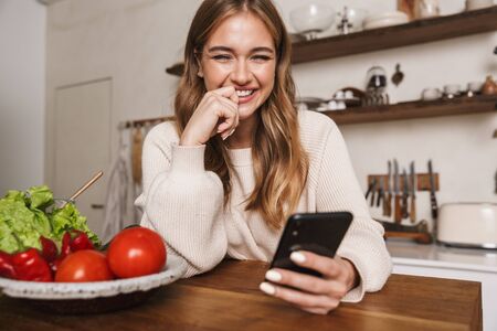 Image of joyful caucasian woman wearing casual clothes using cellphone while cooking dinner in cozy kitchenの写真素材