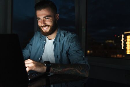 Smiling young man working on laptop computer while sitting at the table indoors at nightの写真素材