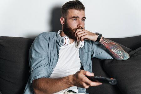 Attractive bored young bearded man relaxing on a couch at home, watching tvの写真素材
