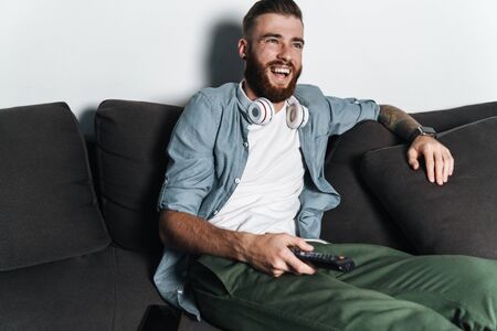Attractive smiling young bearded man relaxing on a couch at home, watching tv, holding remote controlの写真素材
