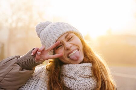 Close up of a cheerful pretty young girl wearing winter jacket and a hat standing on a city street, grimacingの写真素材