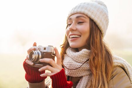 Close up of a cheerful pretty young girl wearing winter jacket and a hat standing on a city street, holding photo cameraの写真素材